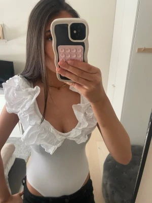 Vit body med volangdetaljer - Snygg vit body med urringning och volanger vid axlarna. Tight passform och ribbat material. 