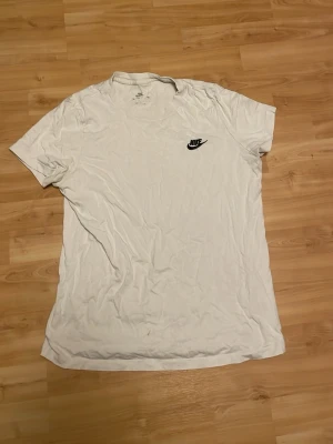 Vit Nike t-shirt med logga - Klassisk vit t-shirt från Nike med svart logga på bröstet. T-shirten har rund halsringning och korta ärmar. Tillverkad i mjuk bomull för en bekväm känsla. Perfekt till vardags för en sportig och enkel stil.