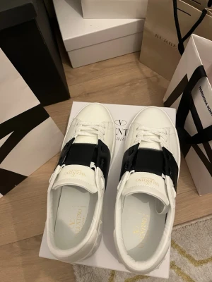 Valentino vita sneakers med svart band - Snygga vita sneakers från Valentino med ett brett svart band över vristen och coola nitar på sulan. Skorna har en stilren design i läder och klassisk låg profil. Perfekta för dig som vill ha en exklusiv och trendig look.