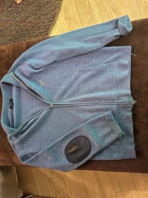 Blå ziptröja med mockadetaljer - Blå stickad ziptröja med ribbad krage och svarta mockadetaljer på armbågarna. Tröjan har två fickor framtill och ribbade muddar vid ärmslut och nederkant. Perfekt för en avslappnad och stilren look.