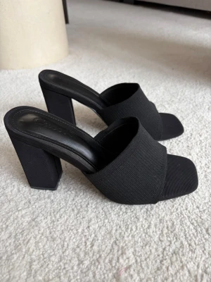 Svarta chunky klackskor - Stilrena svarta klackar med bred ribbad rem över foten och fyrkantig tå. Skorna har chunky blockklack och öppen häl, vilket ger en modern look. Tillverkade i textil och syntetmaterial för en bekväm känsla.
