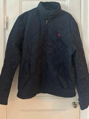 Marinblå quiltad jacka Polo Ralph Lauren - Snygg marinblå quiltad jacka från Polo Ralph Lauren med hög krage, dragkedja och knappstängning. Jackan har två fickor med lock och knapp samt det klassiska röda Polo-logot på bröstet. Perfekt för dig som vill ha en stilren och tidlös look.