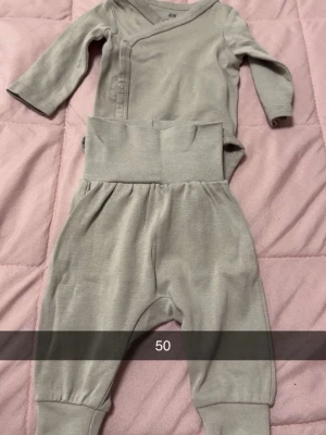 Gråbrun babyset från H&M - Supersött set från H&M med långärmad omlottbody och matchande byxor i mjuk, gråbrun bomull. Bodyn har tryckknappar framtill och byxorna har bred mudd i midjan för extra komfort. Perfekt för små bebisar som vill ha det bekvämt och stilrent.