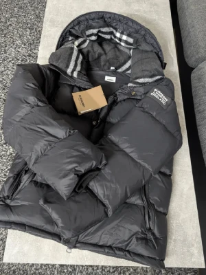 Svart dunjacka från Burberry - Säljer en svart dunjacka från Burberry med klassiskt rutigt foder i huvan. Jackan har dragkedja, tryckknappar och två sidofickor. Oversized modell med hög krage och justerbar huva. Perfekt för kalla vinterdagar och riktigt snygg streetstil.