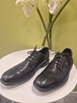 Rieker svarta loafers i läder med snörning 44 - Stilrena svarta loafers i äkta läder med klassisk snörning och diskret logodetalj på sidan. Skorna har en låg klack och rundad tå, perfekta för en clean och dressad look. Snygga detaljer i brunt runt snörningen och sula.