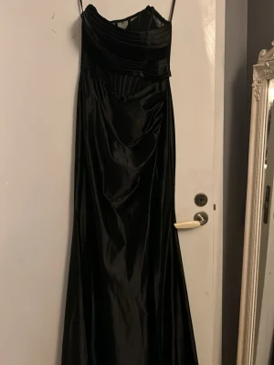 Svart långklänning med axelband - Elegant svart långklänning med draperad överdel. Klänningen har en glansig finish och är figurnära upptill med ett vackert fall. Perfekt för speciella tillfällen som t.ex bal och andra festligheter, kläningen har en korset så det går att justera hur du själv vill ha den , kläningen är i spets upptill och satin nertill , köpt på Shein , använd en gång i några timmar 