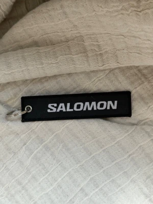 Salomon svart nyckelring - Snygg svart nyckelring från Salomon med vit broderad logga. Rektangulär form och metallring för enkel fastsättning på nycklar eller väska. Perfekt accessoar för dig som gillar sportiga detaljer.
