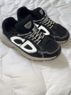 Svarta Dior sneakers med reflekterande detaljer - Snygga svarta Dior sneakers med stora reflekterande CD-detaljer på sidan och vita snören. Skorna har en chunky sula och meshpaneler för extra komfort. Perfekta för dig som vill sticka ut med exklusiv design och streetwear-vibe.