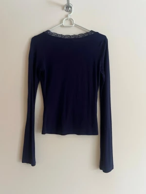 Mörkblå långärmad topp med spets - Mörkblå långärmad topp med v-ringning och fina spetsdetaljer runt halsen. Figurnära passform som framhäver siluetten. Perfekt att styla med jeans eller kjol för en trendig look.