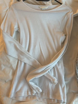 Vit ribbad långärmad - Säljer en enkel och stilren vit ribbad långärmad topp från H&M. Toppen har en rund halsringning och är figurnära i modellen, perfekt att matcha med jeans eller kjol. Materialet är mjukt och stretchigt för en bekväm passform.