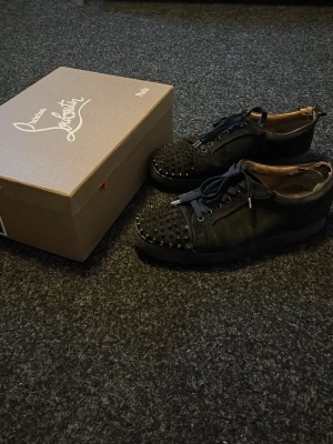 Svarta Louboutin sneakers med nitar - Säljer ett par svarta sneakers från Christian Louboutin med coola nitar på tån och klassisk röd sula. Skorna har snörning. Perfekta för dig som vill sticka ut med exklusiv streetstyle. Dem är ganska slitna går putsa upp med ork och saknas en påse med extra snören men snörerna mår bra annars saknas en spik på en sko.