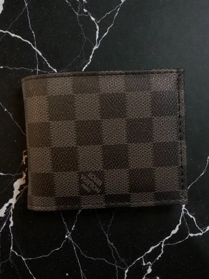 Lton plånbok i damier-mönster - Snygg brun och svart plånbok från Ltton med klassiskt damier-mönster. Plånboken är i läder och har flera kortfack samt ett fönsterfack på insidan. Perfekt för dig som gillar stilrena accessoarer med lyxig känsla.