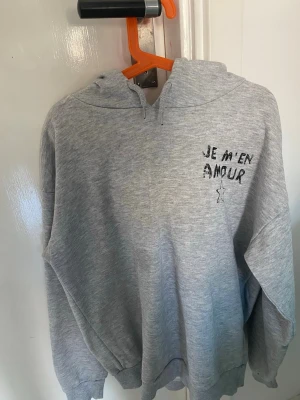 Grå hoodie med tryck 'JE M'EN AMOUR' - Säljer en grå hoodie med svart texttryck 'JE M'EN AMOUR' och en stjärna på bröstet. Hoodien har huva och ribbade muddar vid ärmslut och nederkant. Perfekt för en chill och avslappnad stil. Materialet känns mjukt och bekvämt.