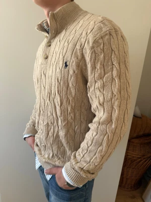 Beige kabelstickad tröja Polo Ralph Lauren - Kabelstickad beige tröja från Polo Ralph Lauren med hög krage och fyra knappar. Tröjan har ribbade muddar och den klassiska svarta logotypen broderad på bröstet. Storlek XL i bra skick. Skjortan under finns också på min plick