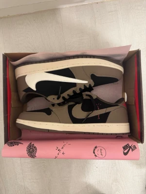 Nike Air Jordan 1 Low Travis Scott - Nike Air Jordan 1 Low Travis Scott med bruna och svarta paneler, vit omvänd Swoosh och röda broderade detaljer på hälen. Skorna har svart snörning, beige sula och är tillverkade i mocka och skinn. Ikonisk collab med Travis Scott och snygg kontrast mellan färgerna.