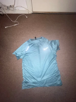 Blå Nike löpartröja - Snygg blå kortärmad löpartröja från Nike med reflekterande logga och texten 'RUNNING' på bröstet. Tillverkad i ett lätt och ventilerande funktionsmaterial som passar perfekt för träning och löpning.