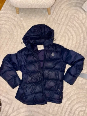 Mörkblå dunjacka från GANT - Snygg mörkblå dunjacka från GANT med huva och dragkedja. Jackan har två framfickor med dragkedja och en puffig, quiltad design som håller dig varm under kalla dagar. Perfekt för vintern och har en klassisk look med diskret logga på bröstet.