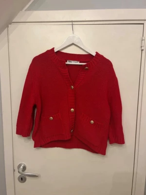 Röd kofta från Zara med guldknappar - Riktigt snygg röd kofta från Zara med trekvartsärmar och dekorativa guldknappar framtill. Koftan har två fickor och ribbade kanter vid hals, ärmslut och nederkant. Perfekt att slänga över en t-shirt för en chill look.