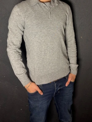 Ljusgrå stickad Filippa K tröja M  - Snygg ljusgrå stickad tröja från Filippa K med klassisk krage och knäppning vid halsen. Tröjan har långa ärmar och ribbade muddar vid ärmslut och nederkant. Perfekt för en stilren och enkel look.