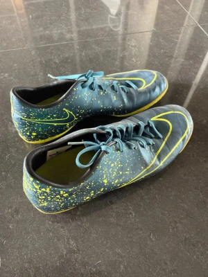 Nike blå-gula fotbollsskor - Säljer ett par Nike fotbollsskor i blått med gula detaljer och stänkdesign. Skorna har snörning, låg profil och är tillverkade i syntetmaterial. Perfekta för dig som vill sticka ut på planen med stilrena och sportiga skor. Fotbollsskor / futsalskor. Skorna säljs inte längre, är ett av Nike mercurial og shoes. Vill du sticka ut på planen, samt vara unik. Är detta för dig!