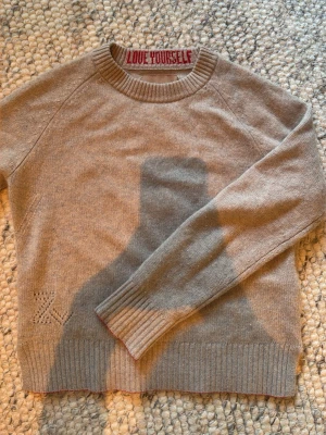 Beige stickad tröja med textdetalj - Beige stickad tröja med rund hals och ribbade muddar. På insidan av kragen står det 'LOVE YOURSELF' i rött och vid nederkanten finns ett diskret hålmönster. Tröjan har en avslappnad passform och är perfekt för lager på lager.