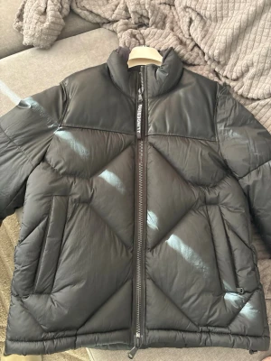 Svart pufferjacka från J.Lindeberg - Säljer en svart pufferjacka från J.Lindeberg med hög krage och dragkedja framtill. Jackan har två stora snedställda fickor och coola sömdetaljer i geometriskt mönster. Materialet är glansigt och ger en modern vibe. Perfekt för kalla dagar. Jackan är helt ny och oanvänd med pislapp på.