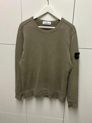 Stone Island Sweatshirt  - Snygg olivgrön sweatshirt från Stone Island med klassisk rund hals och ribbade muddar. Tröjan har den ikoniska loggan på vänster ärm och är tillverkad i mjuk bomull. Perfekt för en avslappnad och trendig look.