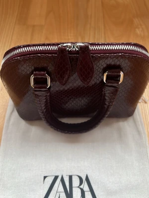 Vinröd handväska i ormmönstrat från Zara - Snygg vinröd handväska från Zara i ormmönstrat konstläder. Väskan har både handtag och avtagbar axelrem samt silverfärgade detaljer och dragkedja. Perfekt för dig som vill ha en trendig accessoar med plats för det viktigaste.