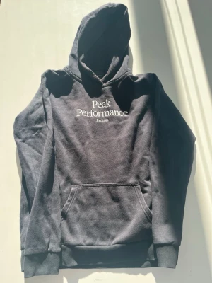 Svart hoodie Peak Performance - Snygg svart hoodie från Peak Performance med vit logga och text på bröstet. Klassisk känguruficka framtill och ribbade muddar vid ärmar och nederkant. Tillverkad i mjukt bomullsmaterial och har en stor huva för extra komfort. Jag har använt den typ 2 gånger och man kan inte se att den är använd. Ny kostar 800 