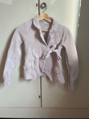  stickad kappa med knytskärp - Snygg beige stickad kappa med grovstickad struktur och knytskärp i midjan. Kappan har två stora fickor framtill och en hög krage. Perfekt för kyliga dagar när du vill vara både varm och stilren.