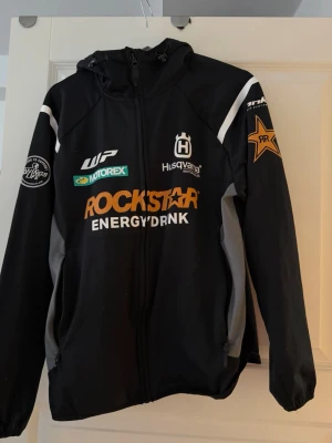 Rockstar Husqvarna svart hoodie - Svart hoodie med dragkedja från Husqvarna Motorcycles, dekorerad med stora Rockstar Energy Drink-loggor och flera motorsportmärken. Jackan har huva, vita och grå detaljer samt tryck på ärmar och bröst. Perfekt för dig som gillar motocross och streetwear.
