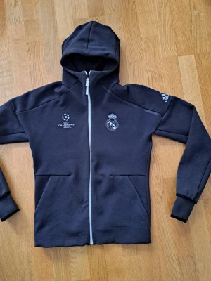 Real Madrid jacka/hoodie med dragkedja - Svart jacka/hoodie från Adidas med Real Madrid och UEFA Champions League-logga på bröstet, perfekt för dig som älskar fotboll och vill ha en sportig look.