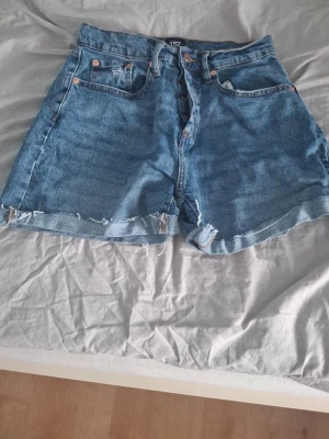 Blå högmidjade jeansshorts - Säljer ett par blå jeansshorts med hög midja och knappar i gylfen. Shortsen har råa kanter nertill och klassiska fem fickor. Perfekta för sommaren och ger en avslappnad vintagekänsla.