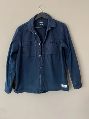 Mörkblå overshirt från Name It - Stilren mörkblå overshirt från Name It med två bröstfickor och knäppning framtill. Skjortan har klassisk krage, långa ärmar och en liten vit patch nertill. Tillverkad i mjukt bomullstyg som ger en avslappnad look.