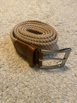 Flätat bälte - Grish beige flätat bälte med bruna skinn-detaljer och silverfärgat spänne. Perfekt accessoar för att lyfta din outfit med en stilren touch. Bältet har en klassisk design och passar till många olika jeans