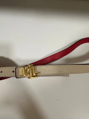 Beige och rött bälte från Ralph Lauren - Snyggt smalt bälte från Ralph Lauren med beige utsida och röd insida. Bältet har ett elegant guldspänne med RL-logga och är gjort i präglat skinn. Perfekt för att lyfta vilken outfit som helst. Ps bältet är i storlek S men funkar även till xs då jag har gjort ett extra hål som visas på bild nr3 och 5!🤍❤️