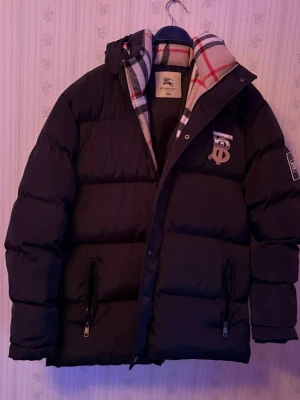 Svart dunjacka från Burberry - Säljer en svart dunjacka från Burberry med klassiskt rutigt foder i beige, rött och svart. Jackan har dragkedjor på fickorna, stor huva och Burberry-logga på bröstet. Perfekt för kalla vinterdagar och riktigt snygg detalj med fodret.