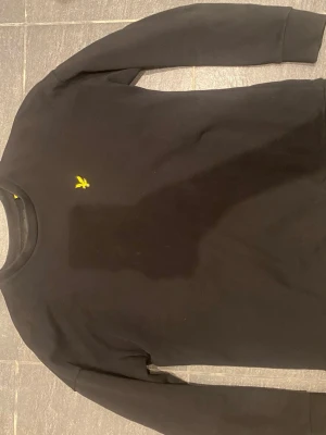 Svart långärmad topp från Lyle & Scott - Klassisk svart långärmad topp från Lyle & Scott med gul broderad logga på bröstet. Tillverkad i mjuk bomull för en bekväm känsla. Perfekt till jeans eller kjol för en clean och stilren look.