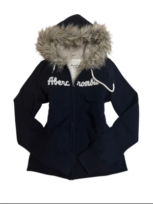 Vintage Marinblå hoodie Abercrombie & Fitch - Mysig marinblå hoodie från Abercrombie & Fitch med dragkedja och stor huva fodrad med fuskpäls. Broderad logga i vitt över bröstet och fickor framtill. Perfekt för dig som vill ha en varm och snygg hoodie med streetkänsla.