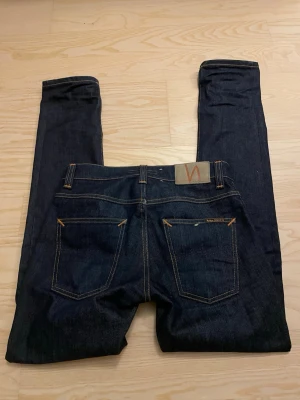 Mörkblå jeans från Nudie Jeans - Säljer ett par mörkblå jeans från Nudie Jeans med klassisk femficksdesign och smal passform. Jeansen har kontrastsömmar och Nudie-logga på bakfickan. Tillverkade i kraftig denim, perfekta för en stilren och enkel look. Passformen heter grim Tim