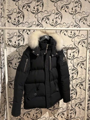 Moose Knuckles herr dunjacka - Svart modell med huva och pälsdetalj. Varm och rejäl jacka med högkvalitativ dunfyllning samt slitstarkt yttertyg.  Jackan är i mycket bra skick – inga synliga skador, fläckar eller slitage. Pälsen är välvårdad och i fint skick. Endast använd ett fåtal gånger.