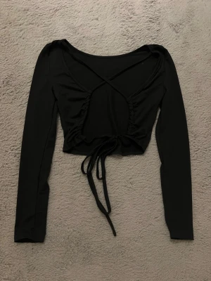 Svart croppad topp med snörning i ryggen - Trendig svart croppad topp med långa ärmar och snygg snörning baktill. Toppen har en omlott-look vid ryggen och är tillverkad i ett mjukt, stretchigt material som sitter tight. Perfekt att styla med höga jeans eller kjol.