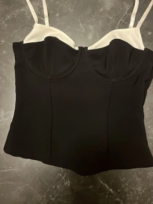 Svart och vit korsettlinne med bygel - Snygg korsettinspirerad topp med svarta kupor och kropp samt vita detaljer upptill och vita justerbara axelband. Toppen har bygel för extra stöd och en figurnära passform. Perfekt att styla med jeans eller kjol för en trendig look.