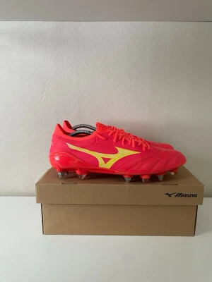 Mizuno neonröda fotbollsskor Elite - Säljer ett par neonröda fotbollsskor från Mizuno med gula detaljer. Skorna har dobbar för bra grepp på gräs. Aldrig använd, skorna är gjorda av ett skönt och formande läder. Säljer för att dem är för små.