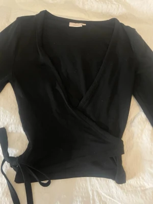 Svart omlottopp med knyt - Snygg svart omlottopp med långa ärmar och knyt i sidan. Använd två gånger men ser helt ny ut. Storlek xs
