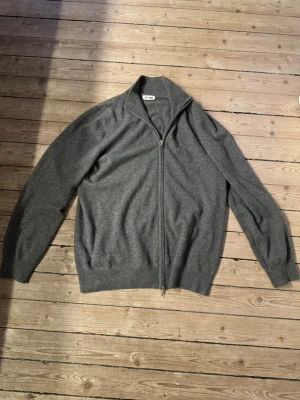 Grå ziptröja i kashmir från Soft Goat - Mysig grå stickad ziptröja från Soft Goat i mjuk kashmir. Tröjan har hög krage, hel dragkedja framtill och ribbade muddar vid ärmslut och nederkant. Perfekt för lager på lager och en clean look. Helt ny med tag, 100% Kashmir! Passar XL