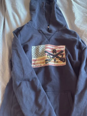 Mörkblå hoodie Polo Jeans Company XS - Mörkblå hoodie från Polo Jeans Company by Ralph Lauren med stor huva och amerikansk flagga med broderade detaljer på bröstet. Tillverkad i mjukt bomullsmaterial och har en klassisk känguruficka framtill. Perfekt för en avslappnad och cool stil.