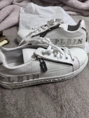 Vita Philipp Plein sneakers med dragkedja - Snygga vita sneakers från Philipp Plein med croco-mönstrad yta, silverfärgade detaljer och dragkedjor på sidorna. Logotypen 'PLEIN' i metall på sidan och nitar längs sulan ger en lyxig vibe. Skorna har vita snören och är låga i modellen.