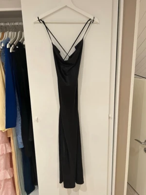 Svart satin långklänning  - Säljer en svart långklänning med tunna axelband som korsas i ryggen. Klänningen har en snygg draperad urringning. 