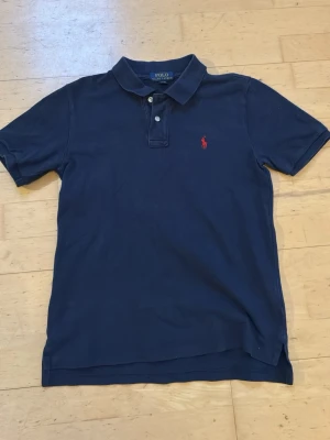 Marinblå pikétröja från Polo Ralph Lauren - Klassisk marinblå pikétröja från Polo Ralph Lauren med röd broderad logga på bröstet. Tröjan har krage, knappar framtill och korta ärmar. Perfekt basic-plagg med snygg passform och tidlös stil.
